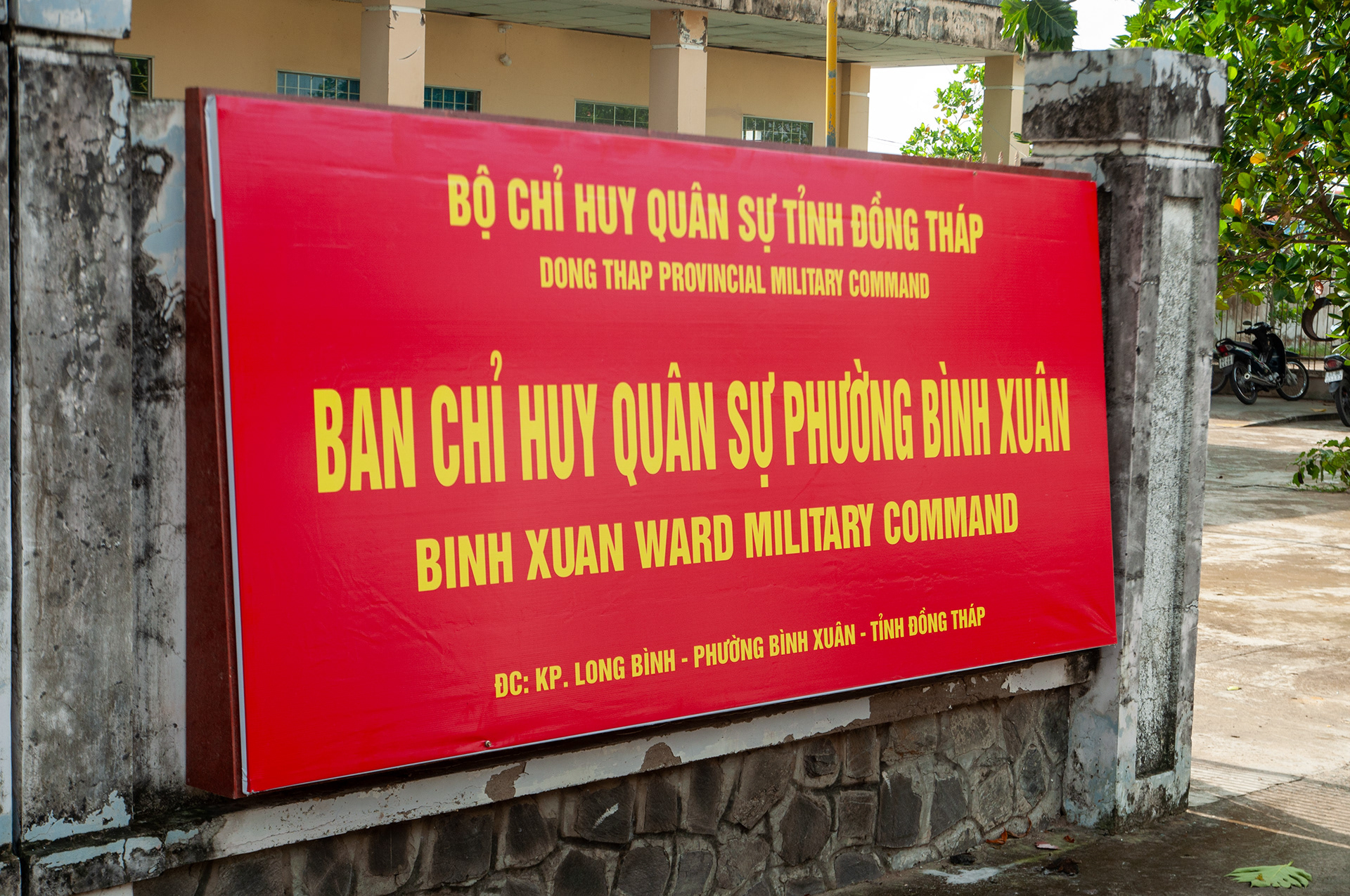 Biển hiệu Ban chỉ huy Quân sự Phường Bình Xuân thuộc Bộ chỉ huy Quân sự tỉnh Đồng Tháp bằng bạt hiflex màu đỏ cờ, chữ màu vàng, lắp cố định giữa hai trụ cổng bê tông cũ của cơ quan.