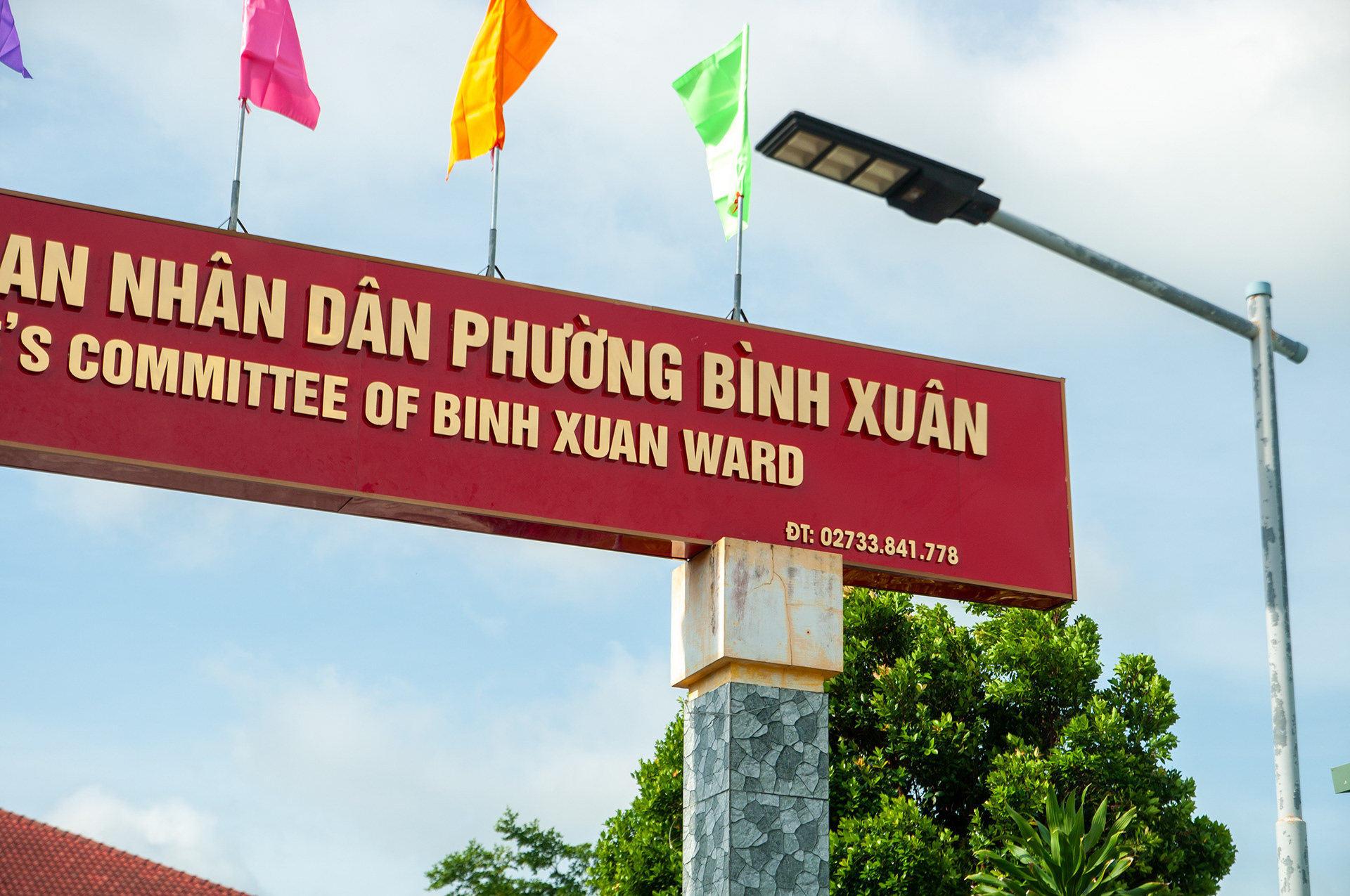 Biển hiệu ngang màu đỏ chữ vàng của Ủy ban Nhân dân Phường Bình Xuân trên trụ đá ngoài trời.